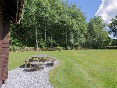 An outdoor area with a round wooden picnic table on gravel beside a lawn and trees at Moyle Lodge in Dalbeattie