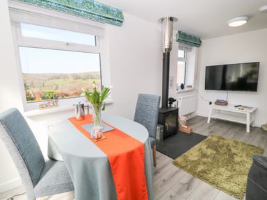 A dining area with a table and two chairs next to a window and a living room with a wood stove a television and a green rug at The View in Cardinham