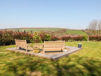 An outdoor wooden deck with benches and potted plants on a grassy lawn with hills in the background at The View in Cardinham