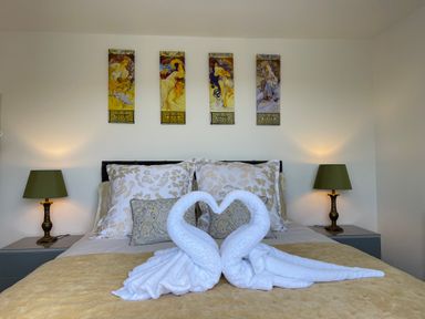 A bedroom with a bed featuring floral pillows towels folded into swans on a beige cover and green lamps on side tables at The View in Cardinham