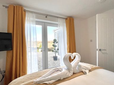 A bedroom with two towel swans on a bed near glass doors with yellow curtains at The View in Cardinham