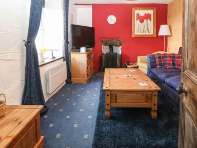 A living room with blue patterned carpet a wooden coffee table a television on a wooden stand a red accent wall with a clock and flower painting at Bod Feurig Penygroes