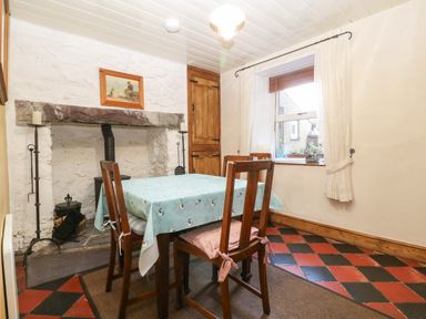 A dining room with a wooden table covered with a blue tablecloth and four chairs near a fireplace with candles and a window with white curtains at Bod Feurig in Penygroes