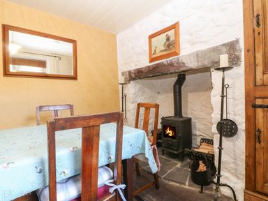 A dining area with wooden chairs around a table covered with a patterned tablecloth and a wood-burning stove in a stone fireplace at Bod Feurig in Penygroes