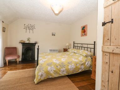A bedroom with a bed yellow floral bedding an armchair a small black fireplace and wooden door at Bod Feurig in Penygroes