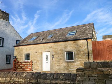 A stone cottage with a white door and two small windows behind a stone wall at Stable Cottage in Hallbankgate