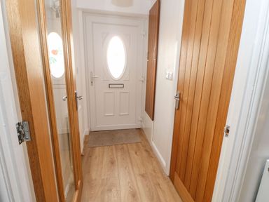A hallway with a white door with an oval window a wooden floor a carpet a wooden door and a tall mirror at Stable Cottage in Hallbankgate
