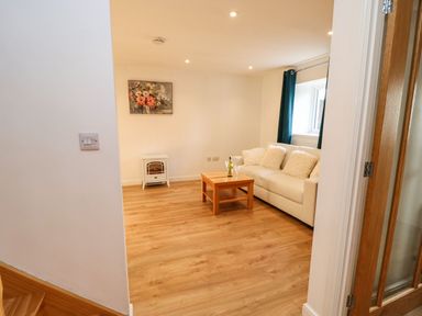 A living room with a white sofa with cushions a wooden coffee table with a bottle and glasses a small heater and a painting on the wall at Stable Cottage in Hallbankgate