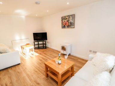 A living room with two white sofas a wooden coffee table with a wine bottle and glasses a small tv on a stand and a floral painting on the wall at Stable Cottage in Hallbankgate