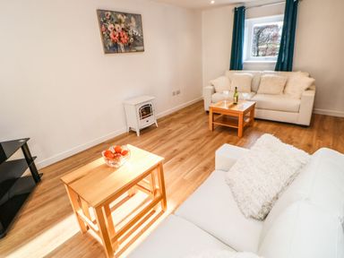 A sitting room with two white sofas wooden tables a black shelving unit and a window with blue curtains at Stable Cottage in Hallbankgate