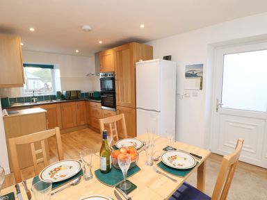 A kitchen with wooden cabinets a white refrigerator a dining table set with plates glasses and a bowl of fruit at Stable Cottage in Hallbankgate