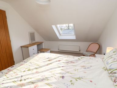 A bedroom with a bed covered with floral bedding a wooden door a white dresser with a mirror a stool a chair and a skylight window at Stable Cottage in Hallbankgate