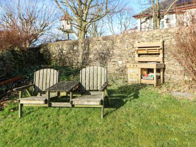 A garden with two wooden chairs and a wooden table between them near a stone wall with a wooden play kitchen next to the wall at Stable Cottage in Hallbankgate