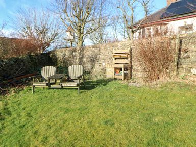 A garden with two wooden chairs and a table on grass near a stone wall and a wooden play kitchen at Stable Cottage in Hallbankgate