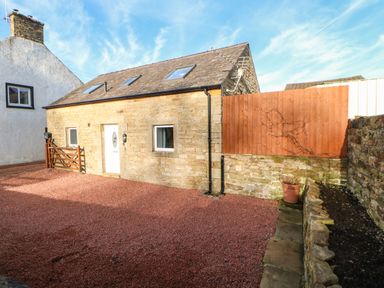 A stone cottage with white door and windows surrounded by red gravel and wooden fence at Stable Cottage in Hallbankgate