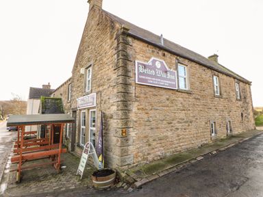An exterior view of a stone building with signs for Belted Will Inn and a wooden bench area at Stable Cottage in Hallbankgate