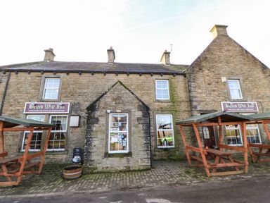 The exterior of a stone building with signs for Belted Will Inn and wooden picnic benches outside at Stable Cottage in Hallbankgate