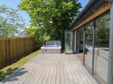 An outdoor deck area with a hot tub next to a glass sliding door showing a living room with a sofa at The Nest Brython in Tenby