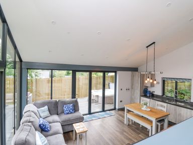 An open plan living room and kitchen with gray sofa, wooden dining table, and sliding glass doors at The Nest Brython in Tenby