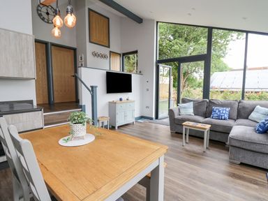 A living room with a grey sofa a wooden dining table and large windows showing outside at The Nest Brython in Tenby