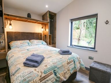 A bedroom with a bed, bedside shelves, window showing trees, and a gray storage bench at The Nest Brython in Tenby