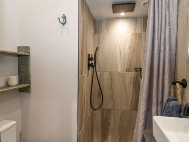 A bathroom with a tiled walk-in shower a black showerhead a gray shower curtain wooden shelves and a sink with blue towels at The Nest Brython in Tenby