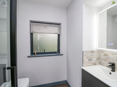 A bathroom with a window featuring a bird patterned blind a modern sink with tiled backsplash and a mirror with lights at The Nest Brython in Tenby