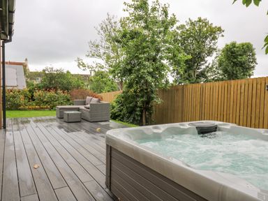 An outdoor deck with a hot tub and grey patio furniture next to a wooden fence and trees at The Nest Brython in Tenby
