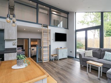A living room with a gray sofa a small wooden table a wall-mounted TV and large windows at Cornerstones Brython in Tenby