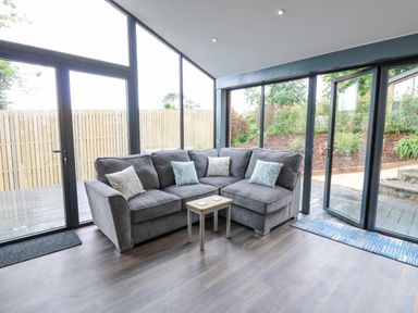 A corner seating area with a grey sectional sofa and small wooden table in a room with large glass windows and doors at Cornerstones Brython in Tenby