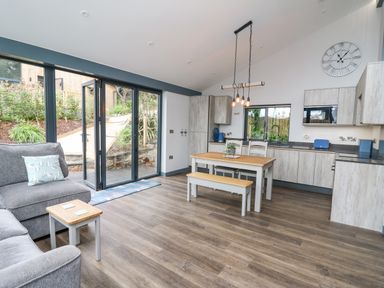 A living area with a gray sofa and pillow a small wooden table a wooden dining table with chairs and bench kitchen cabinets and appliances large glass doors opening to a garden at Cornerstones Brython in Tenby