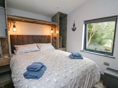 A bedroom with a bed covered in sailboat patterned bedding two sets of blue towels on the bed wooden shelves and hanging lights above the bed a window with a view of trees and a small anchor decoration on the wall at Cornerstones Brython in Tenby