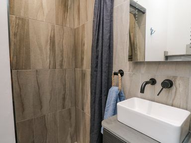A bathroom with a rectangular white sink wall-mounted faucet and beige tiled shower area with a dark shower curtain at Cornerstones Brython in Tenby