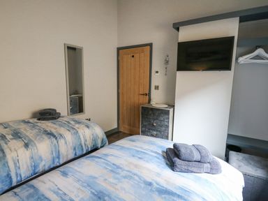 A bedroom with two beds with blue and white bedding towels on beds a wooden door a mirror a chest of drawers a wall-mounted television and an open closet with hangers at Cornerstones Brython in Tenby