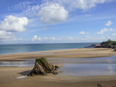 A beach with a large rock formation in the sand and a building on stilts near the shoreline at Cornerstones Brython in Tenby