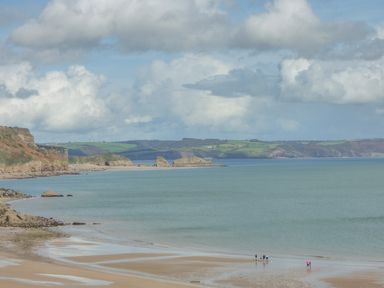 A beach with people walking near the water and rocky cliffs in the background at Cornerstones Brython in Tenby