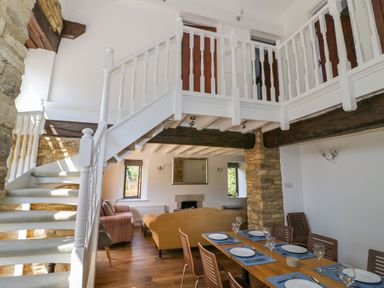 A dining area with a wooden table set with plates and glasses next to a living area with sofas and a fireplace under a white staircase and railing at The Paddock in New Yatt near Witney