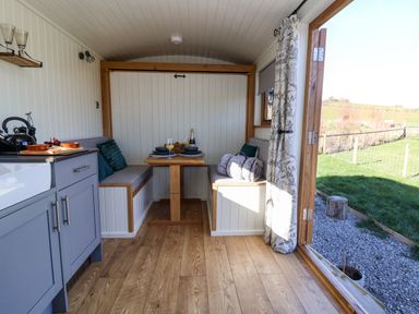 A small dining area with built-in benches and a wooden table next to a kitchen with cabinets and an open door showing an outdoor grass area at Cariad Bach in Gwaenysgor near Prestatyn