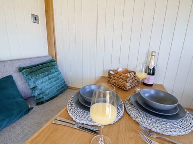 A dining table set for two with gray dinnerware glasses of orange juice a bottle of sparkling wine and a basket of bread with a cushioned bench seat at Cariad Bach in Gwaenysgor near Prestatyn