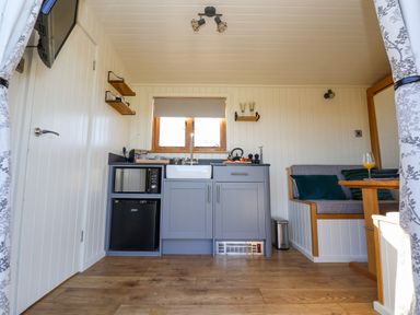 A small kitchen area with gray cabinets a sink microwave and refrigerator next to a wooden dining nook with cushions at Cariad Bach in Gwaenysgor near Prestatyn