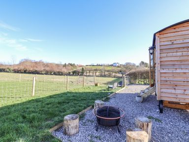 An outdoor area with wooden stools around a fire pit next to a wooden cabin and fenced grassy field at Cariad Bach in Gwaenysgor near Prestatyn