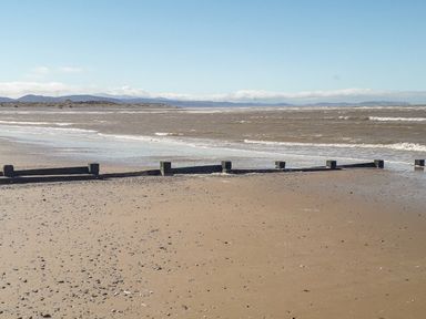A sandy beach with scattered small rocks and wooden groynes with sea waves under a clear sky