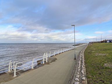A paved walkway along the coast with a white railing and street lamps next to a grassy area by the sea at Cariad Bach in Gwaenysgor near Prestatyn