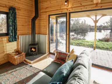 An interior with a wood burning stove a green sofa with cushions a basket with firewood and large glass doors overlooking a pond at Kingfisher in Dunchurch