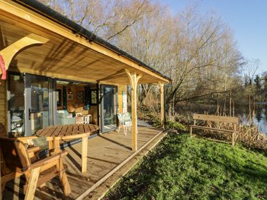 A wooden cabin with a covered porch patio wooden table and chairs and a bench nearby by water at Kingfisher in Dunchurch