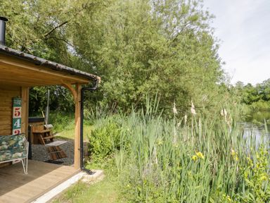 A wooden cabin porch with a patterned chair next to tall reeds and trees by a riverside at Kingfisher in Dunchurch