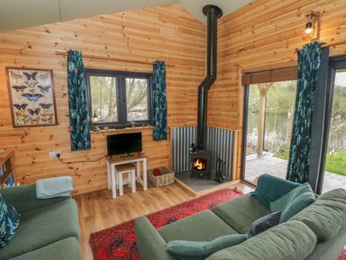 A wooden living room with a wood burning stove green sofas a red rug and a view of a lake at Kingfisher in Dunchurch