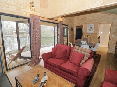 A living area with red sofas a wooden coffee table with duck figurines sliding glass doors leading to a deck with wooden chairs a dining table with white chairs and a wooden interior at Willow in Dunchurch