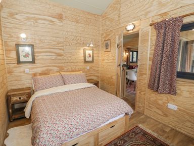 A bedroom with a bed with floral bedding two bedside tables framed pictures a window with curtains and a door into another room at Willow in Dunchurch