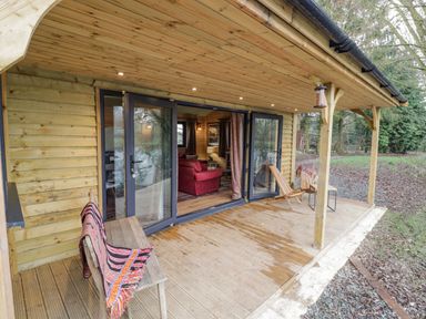 A wooden porch with bench and chair outside a cabin with glass doors showing a living room at Willow in Dunchurch
